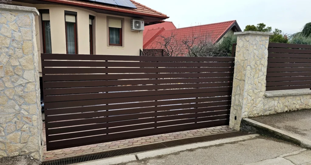 Standard barna alumínium nagykapu terméskő pillérek között, vízszintes lécekkel.