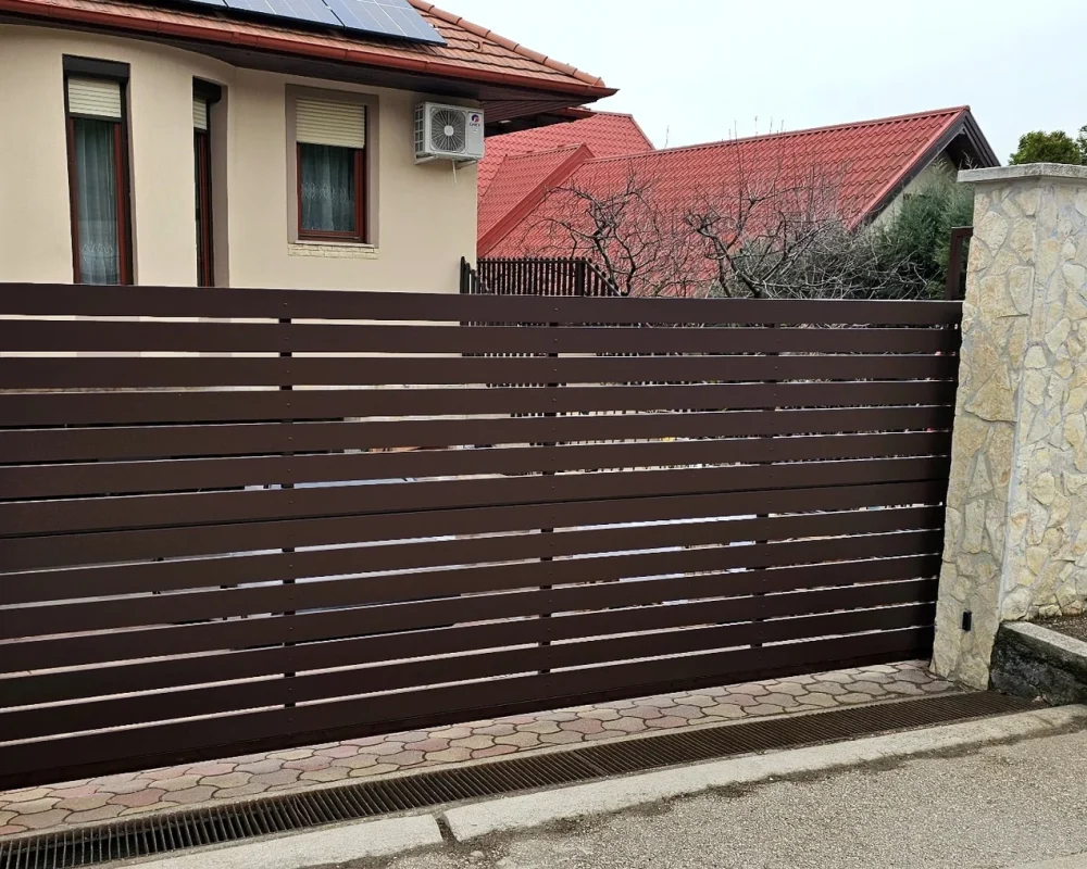 Standard barna alumínium nagykapu terméskő pillérek között, vízszintes lécekkel.